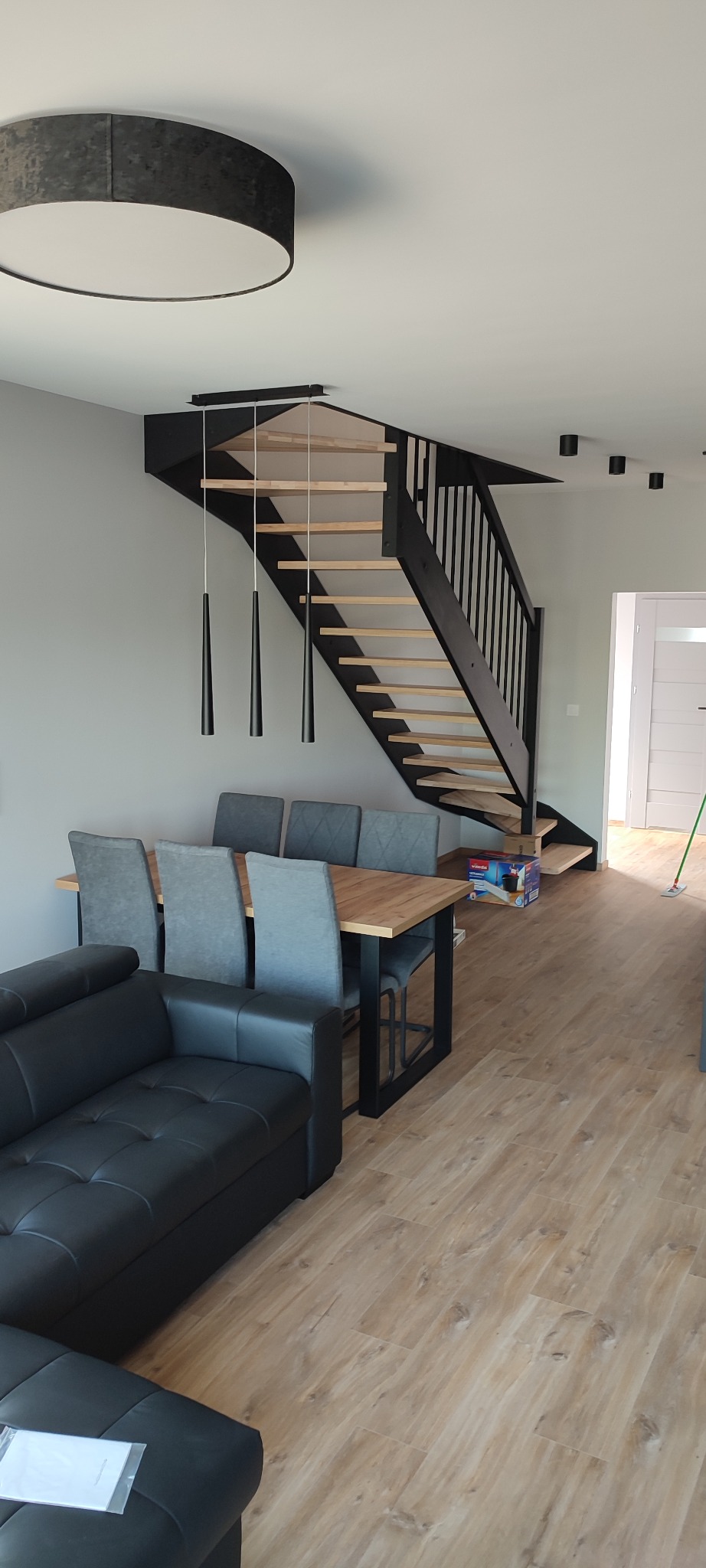 Jasne wnętrze z drewnianymi schodami z czarną balustradą, stołem z szarymi krzesłami i czarną sofą. Wiszące lampy o stożkowym kształcie nad stołem. Podłoga wyłożona panelami imitującymi drewno.