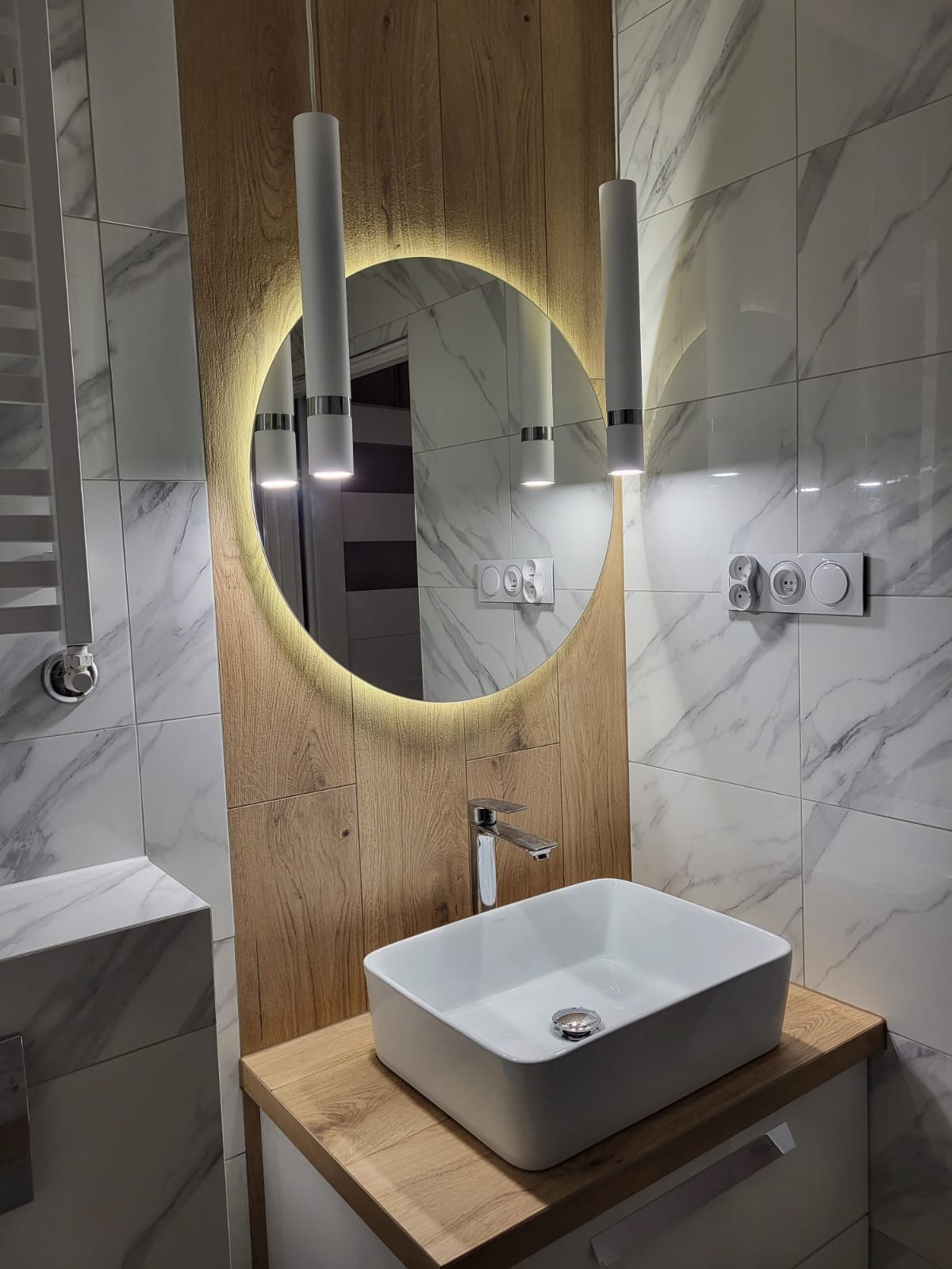 Nowoczesna łazienka z marmurowymi płytkami, okrągłym lustrem z podświetleniem LED, białymi lampami wiszącymi i prostokątną białą umywalką nablatową na blacie imitującym drewno.