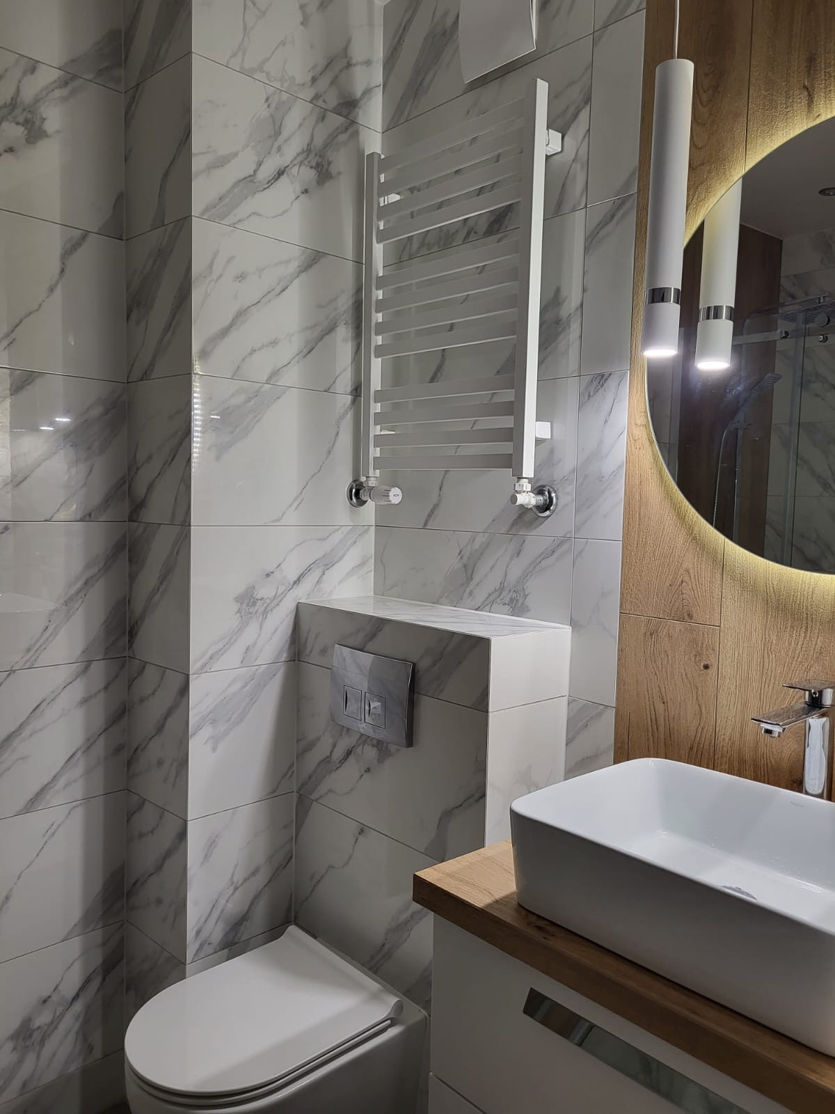Nowoczesna łazienka z marmurowymi płytkami, podwieszaną toaletą, grzejnikiem drabinkowym i okrągłym lustrem z podświetleniem LED.
