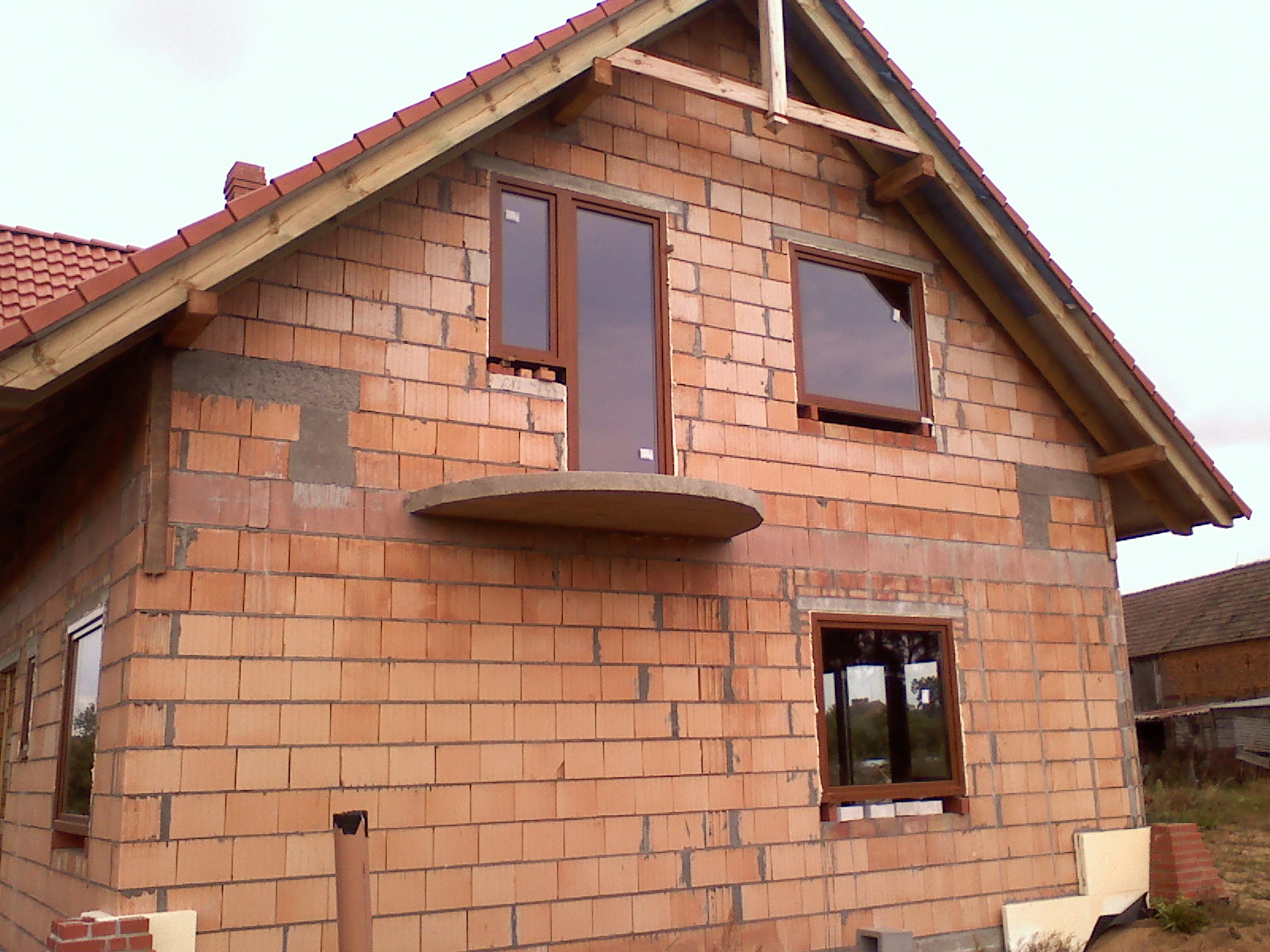 Surowa elewacja domu w trakcie budowy z widocznymi oknami z brązowymi ramami oraz przygotowanym miejscem na balkon.