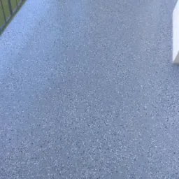 Szary taras z drobnymi, białymi i czarnymi plamkami, widok z góry, z fragmentem metalowej balustrady i drewnianego słupka w tle.