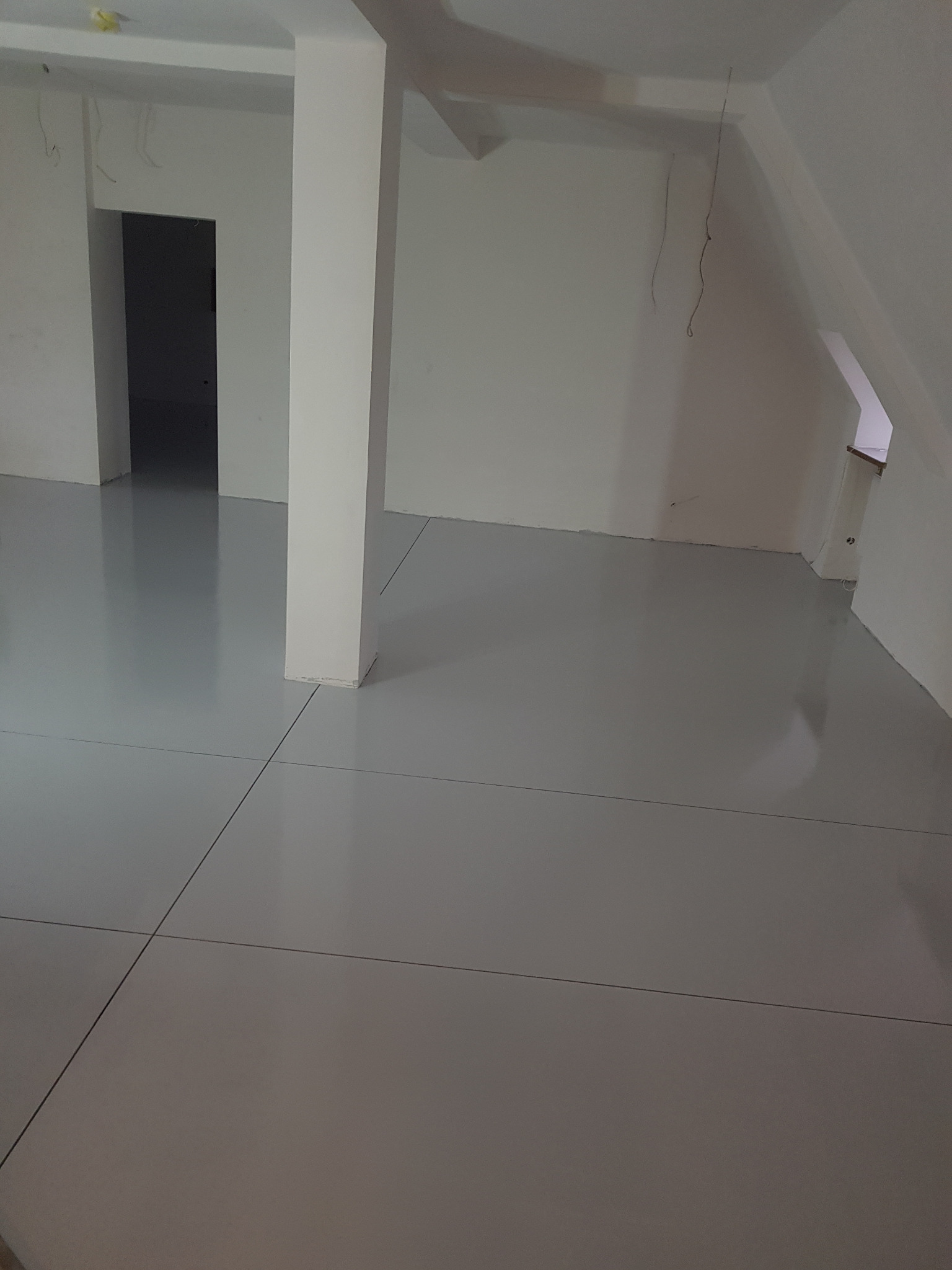 Wykończona podłoga w jasnoszarym odcieniu z widocznymi fugami w przestronnym pomieszczeniu biurowym z kolumną nośną i surowymi kablami zwisającymi z sufitu.