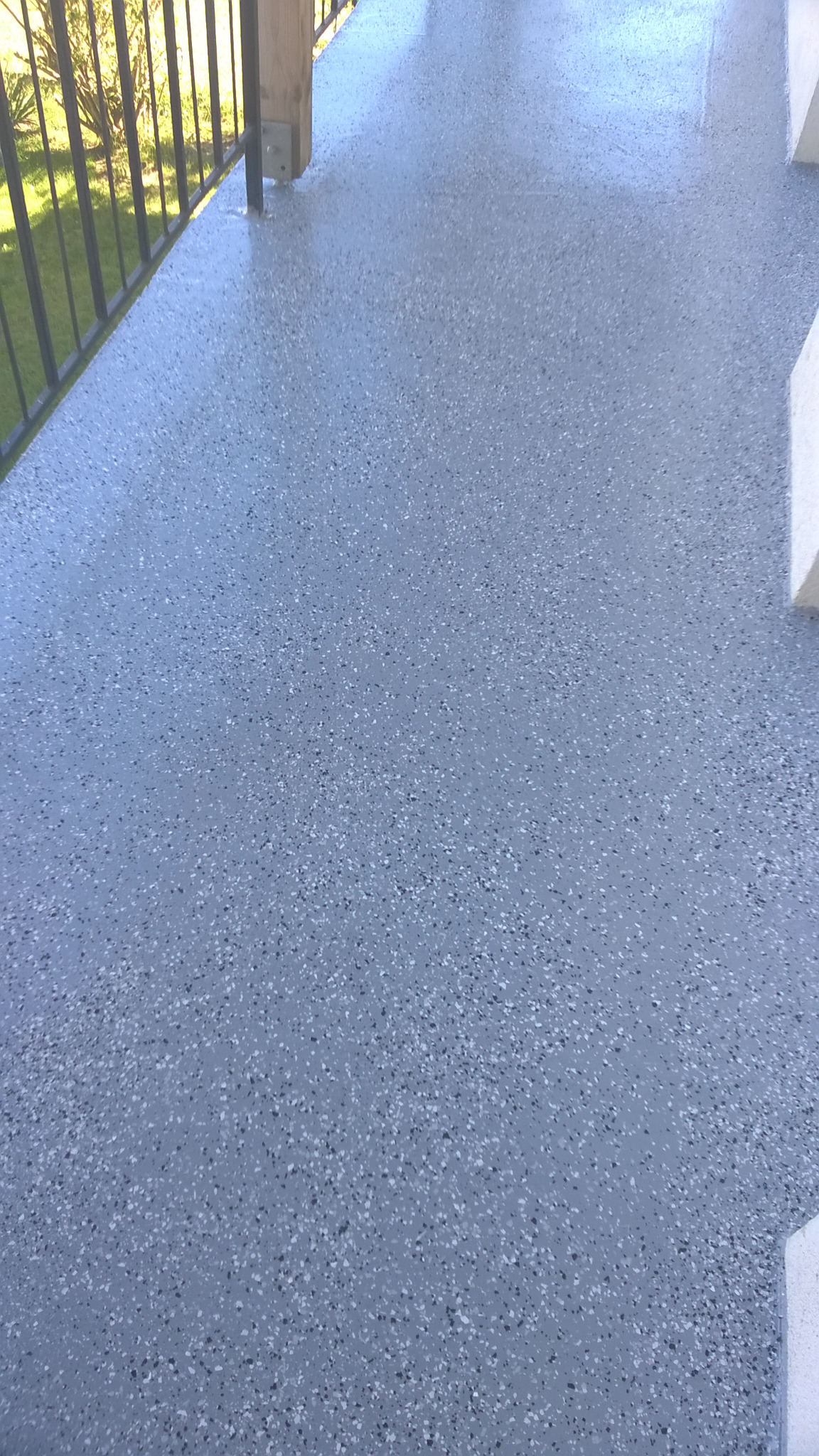 Szary taras z drobnymi, białymi i czarnymi plamkami, widok z góry, z fragmentem metalowej balustrady i drewnianego słupka w tle.