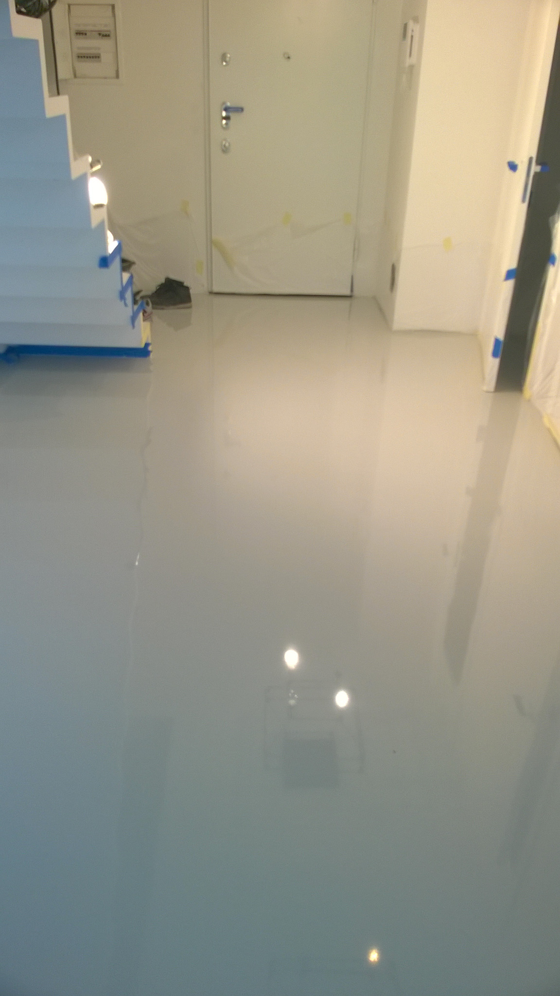 Świeżo wylana, błyszcząca, jasnoszara posadzka żywiczna w trakcie remontu, zabezpieczona folią i taśmą malarską przy drzwiach i schodach.
