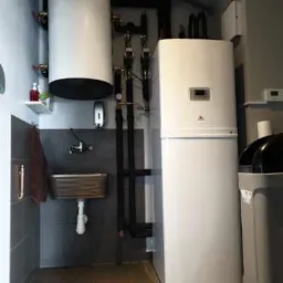 Nowoczesna kotłownia z białym zasobnikiem ciepłej wody i pompą ciepła, widoczna także metalowa umywalka i ciemne rury instalacji.