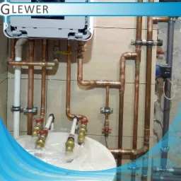 Złożona instalacja rur miedzianych z zaworami, połączona z piecem grzewczym, widoczna na tle płytek ceramicznych.