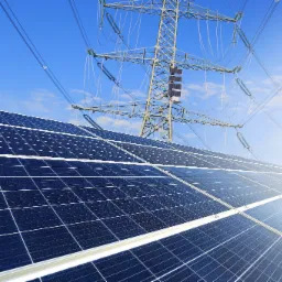 Instalacja paneli słonecznych w Rudzińcu, z widoczną wieżą wysokiego napięcia w tle, pod błękitnym niebem.