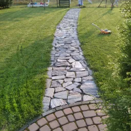 Ścieżka z nieregularnych płyt granitowych prowadząca przez trawnik w kierunku placu zabaw z domkiem, trampoliną i huśtawką, zakończona obrzeżem z kostki brukowej.