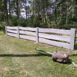 Jasnoszare, drewniane ogrodzenie ranczowe z trzema poprzeczkami na tle lasu, z fragmentem zielonego węża ogrodowego i bryłą ziemi na trawie.