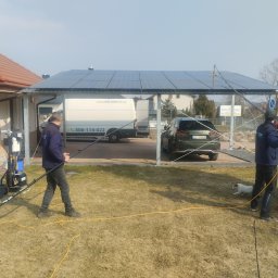 Volton sp. z o.o. - Dwaj pracownicy czyszczą panele fotowoltaiczne na zadaszeniu, używając lanc teleskopowych. Widoczny samochód dostawczy firmy i auto osobowe.