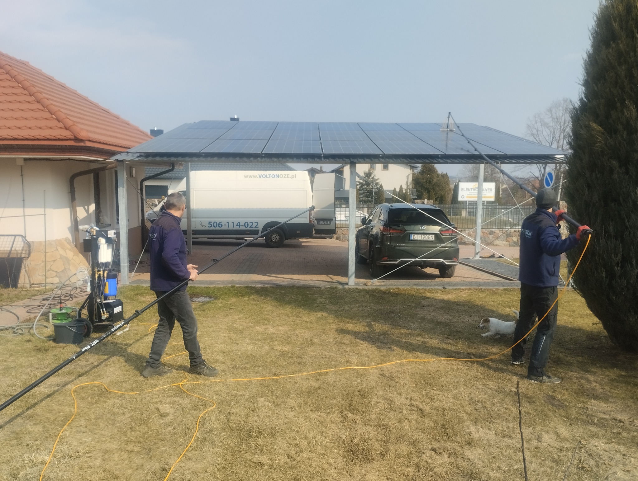 Dwaj pracownicy czyszczą panele fotowoltaiczne na zadaszeniu, używając lanc teleskopowych. Widoczny samochód dostawczy firmy i auto osobowe.