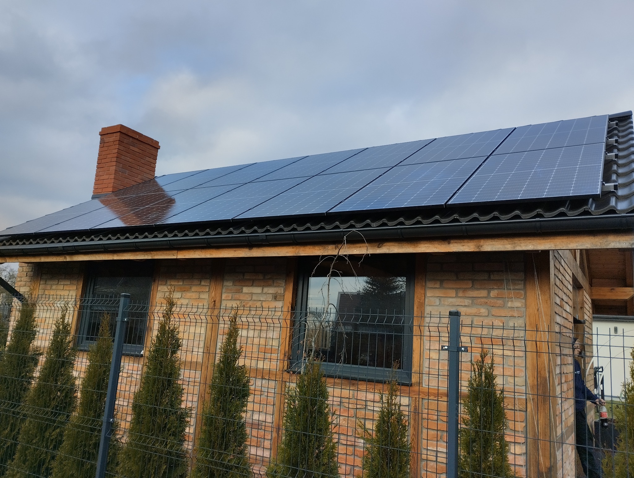 Dom z cegły z panelami fotowoltaicznymi na dachu, widoczny komin z cegły i ogrodzenie z tujami. Pochmurne niebo w tle.