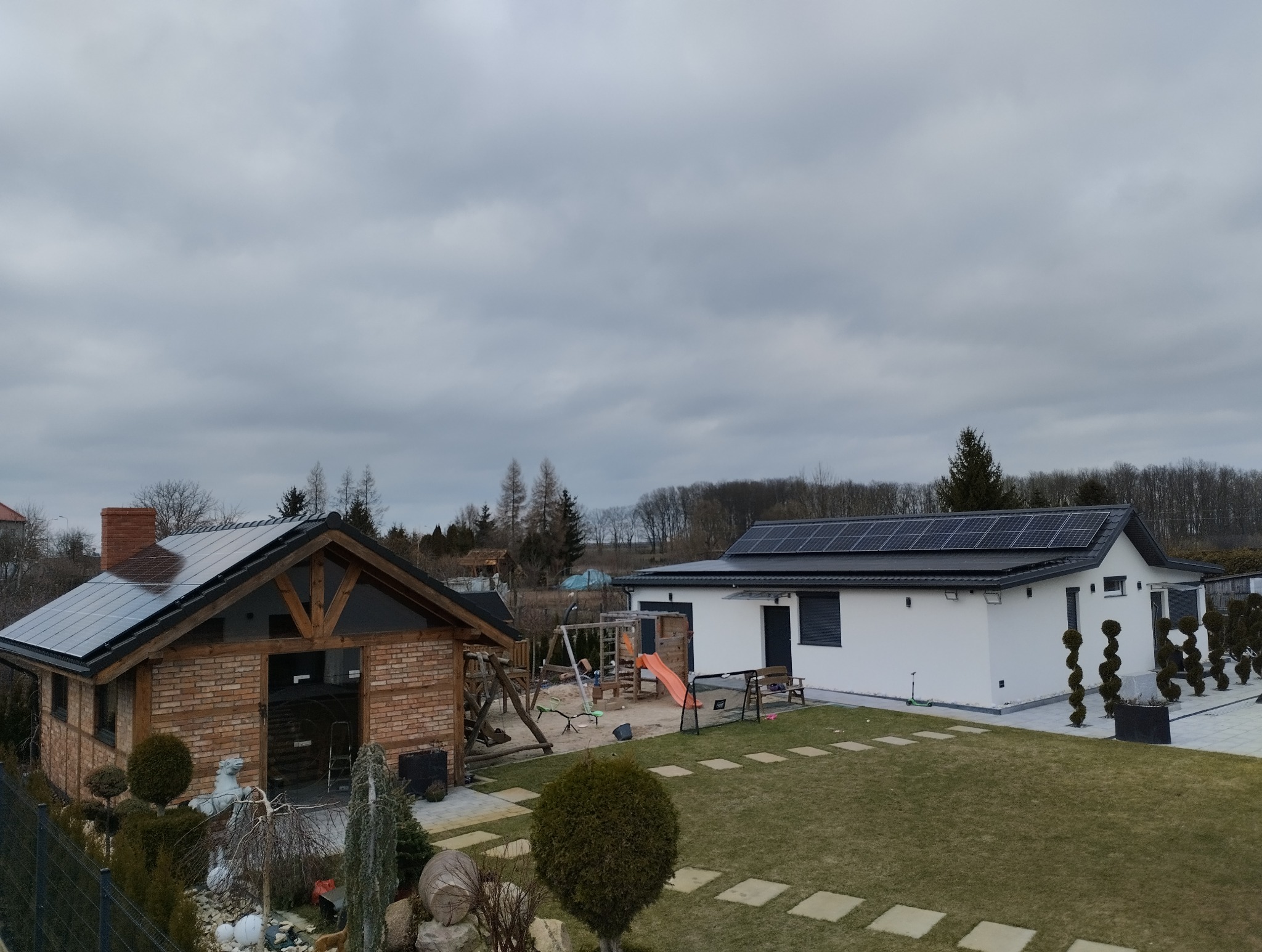 Dwa budynki z panelami słonecznymi na dachach, otoczone zielenią i elementami placu zabaw. Jeden budynek z cegły, drugi biały, nowoczesny. Pochmurne niebo w tle.