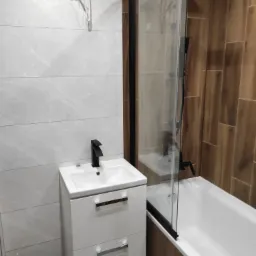 Nowoczesna łazienka po remoncie z białą szafką pod umywalkę z czarną armaturą, kabiną prysznicową z czarnymi profilami i płytkami imitującymi drewno, z widocznymi przewodami elektrycznymi na ścianie.