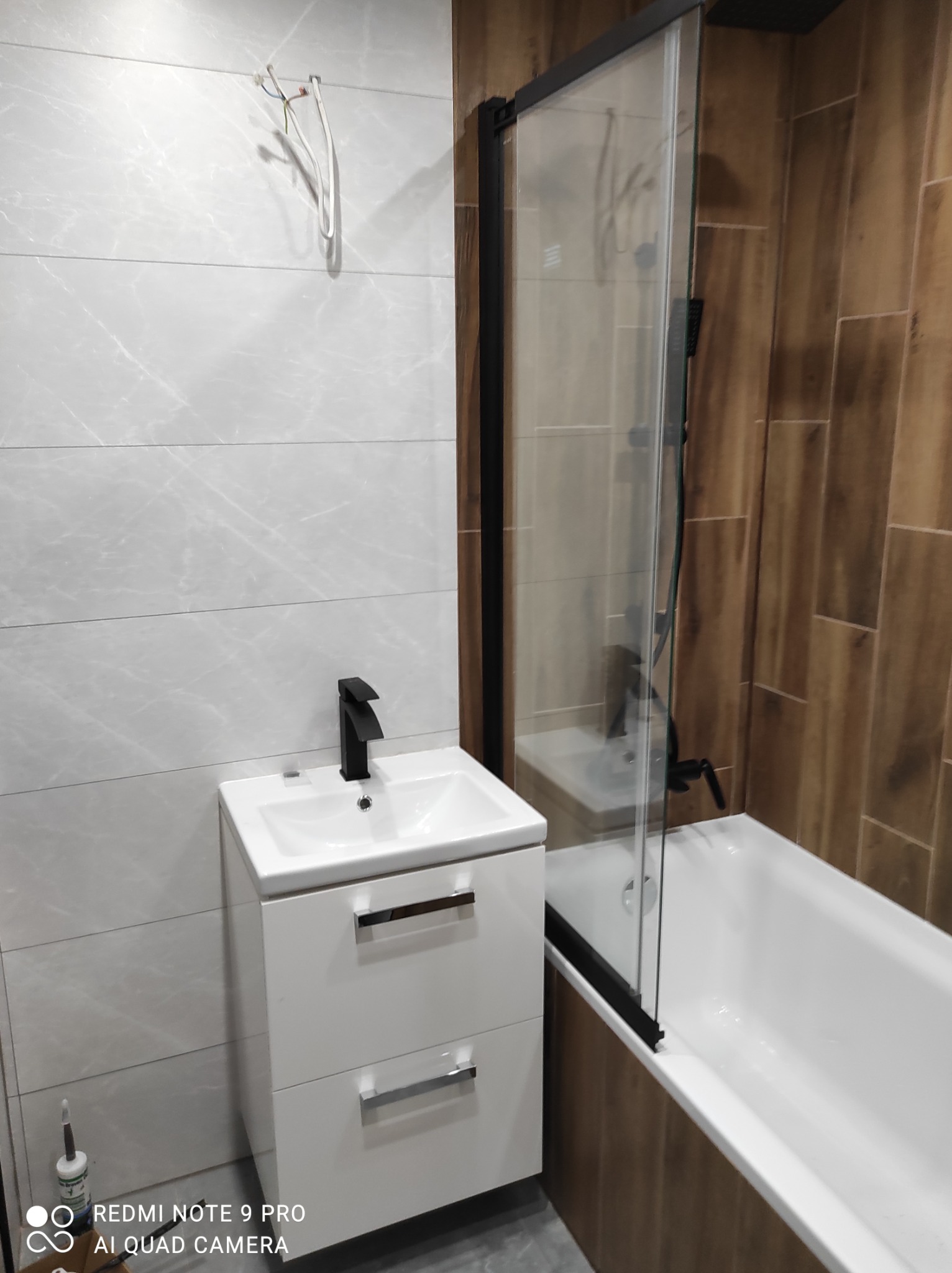 Nowoczesna łazienka po remoncie z białą szafką pod umywalkę z czarną armaturą, kabiną prysznicową z czarnymi profilami i płytkami imitującymi drewno, z widocznymi przewodami elektrycznymi na ścianie.