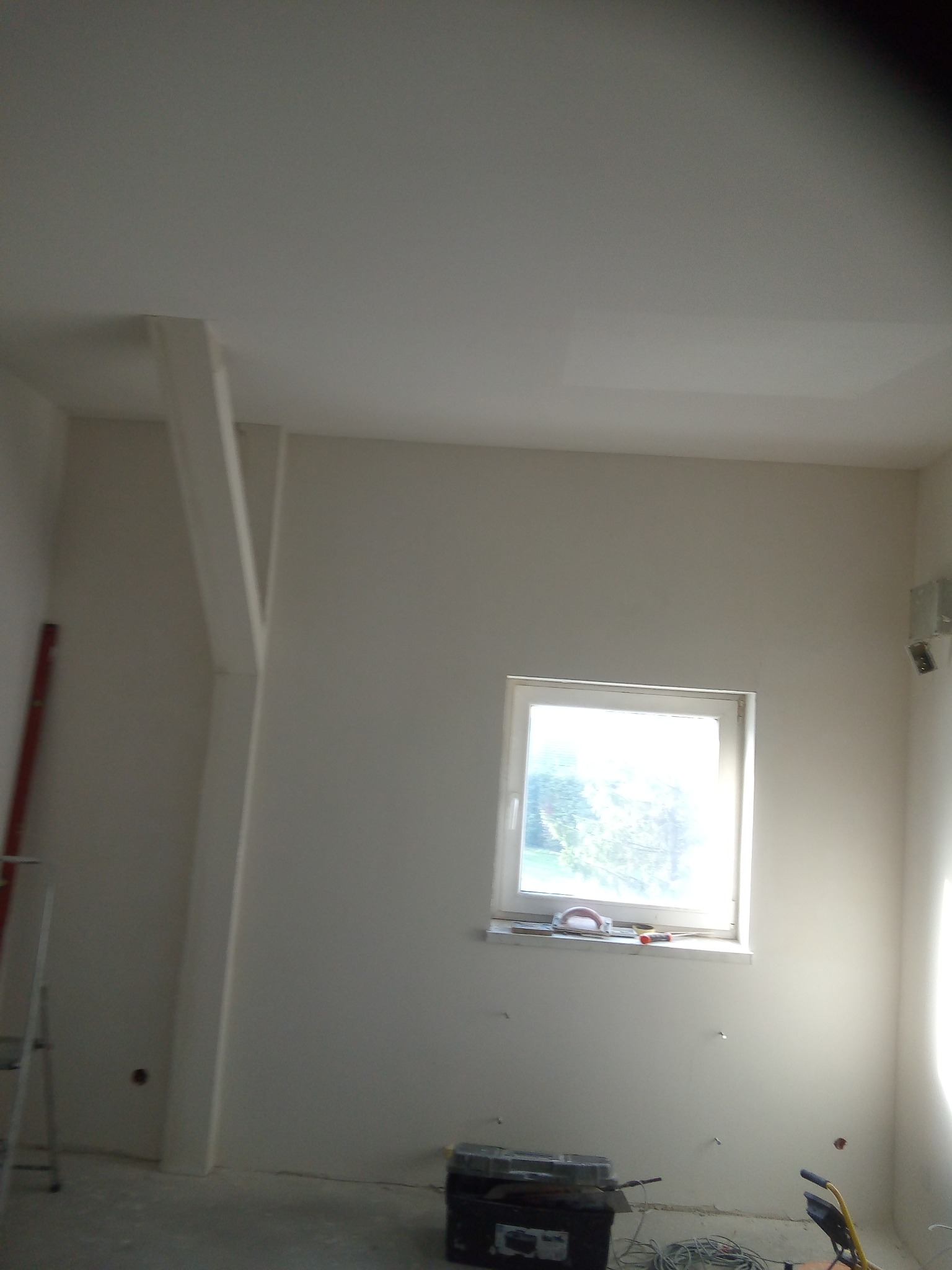 Wnętrze pokoju w trakcie prac wykończeniowych, widoczne narzędzia na parapecie, drabina oparta o ścianę i skrzynka z narzędziami na podłodze.