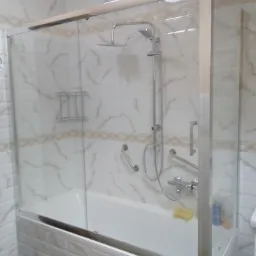 Biała wanna z zamontowaną szklaną kabiną prysznicową z chromowanymi profilami, widoczne uchwyty dla osób z trudnościami w poruszaniu się, ściany wykończone białymi płytkami z delikatnym marmurkowym...