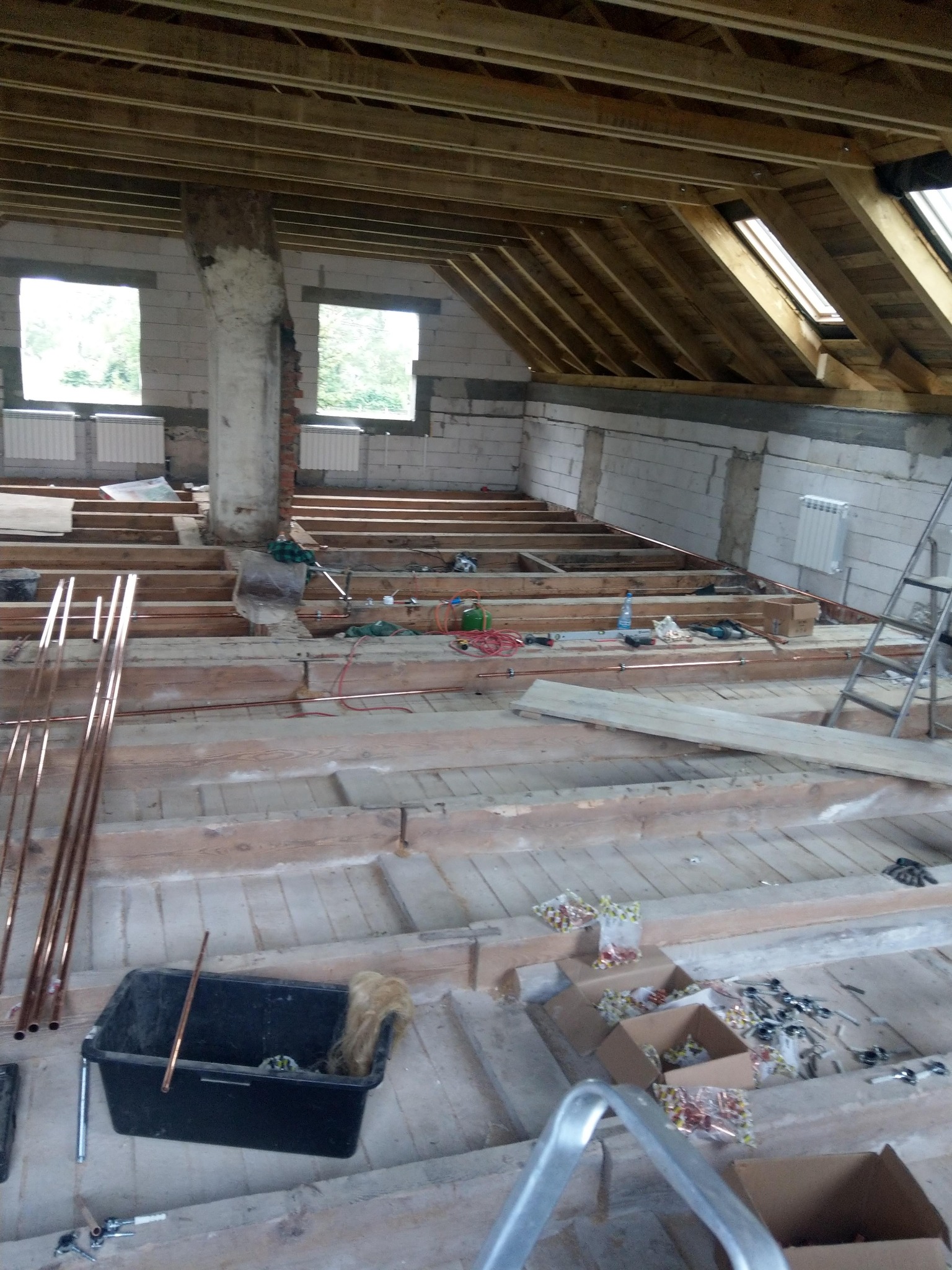 Instalacja grzewcza w trakcie montażu na poddaszu, widoczne rury miedziane, narzędzia i materiały budowlane na drewnianej konstrukcji podłogi, ściany z białego bloczka.