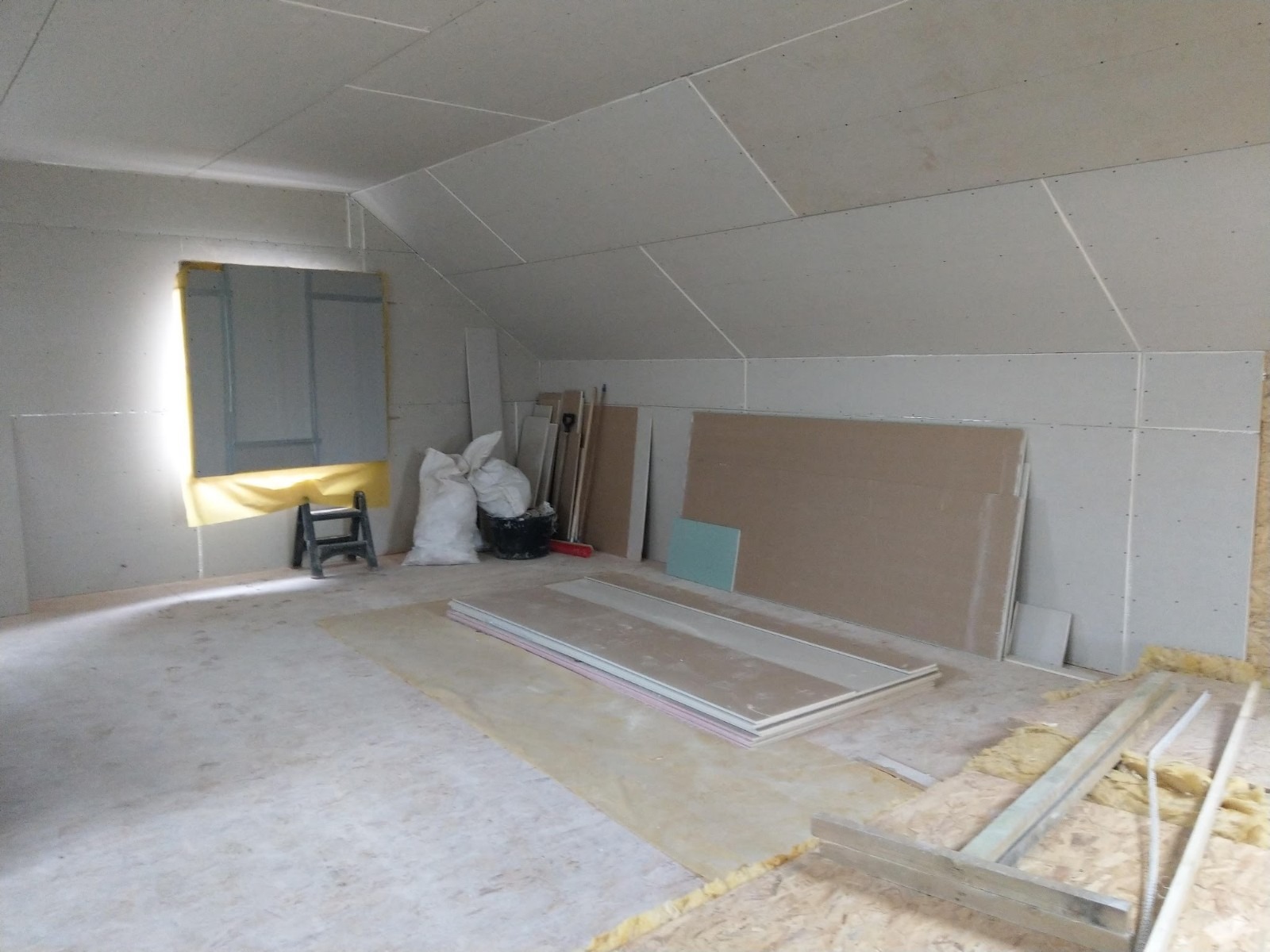 Poddasze w trakcie wykańczania płytami gipsowo-kartonowymi, widoczne przygotowane płyty, drabinka, worki z materiałami budowlanymi oraz zabezpieczone okno.