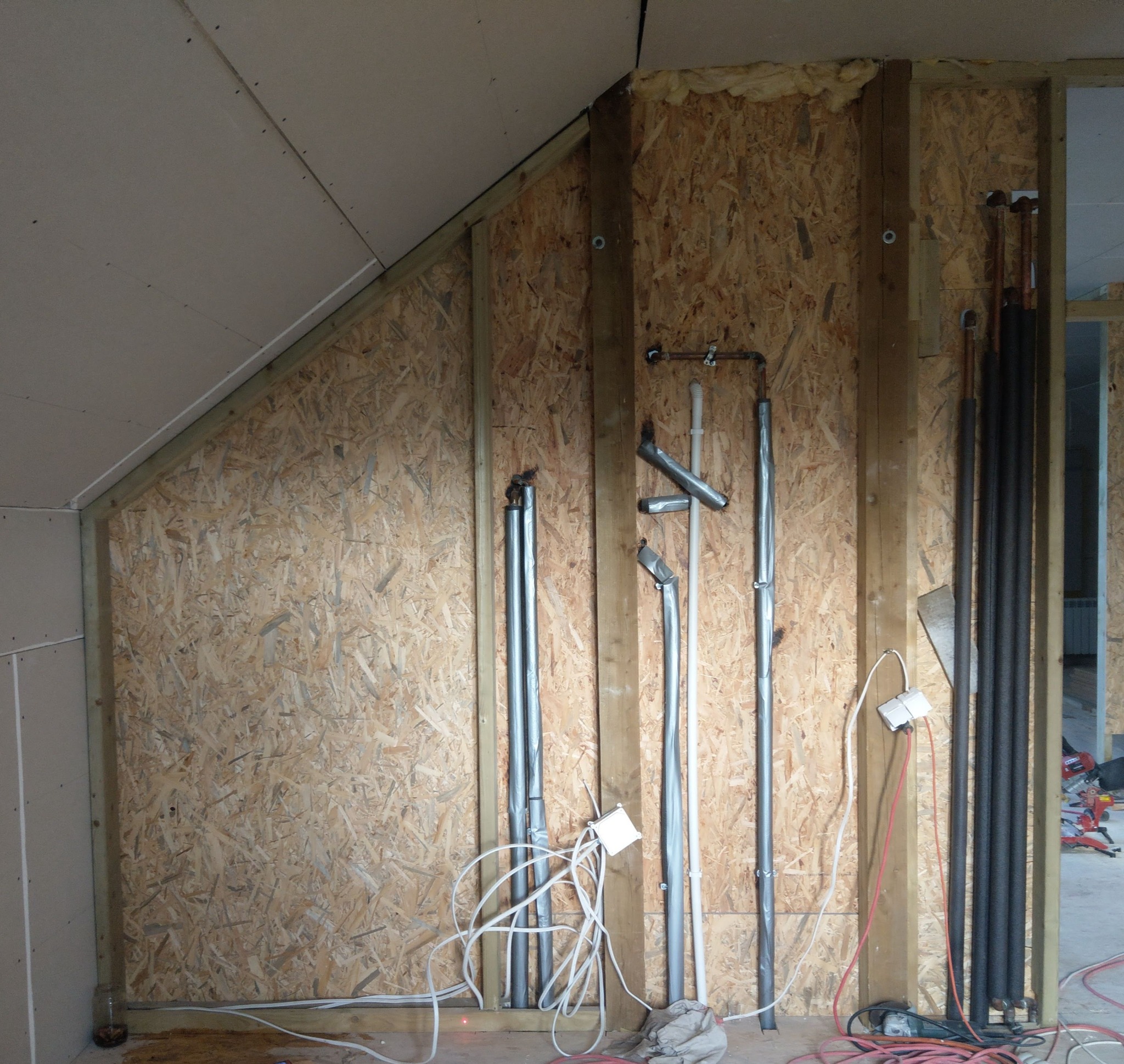 Instalacja rur grzewczych i hydraulicznych w surowym wnętrzu z boazerią, widoczne kable i narzędzia, etap prac wykończeniowych.