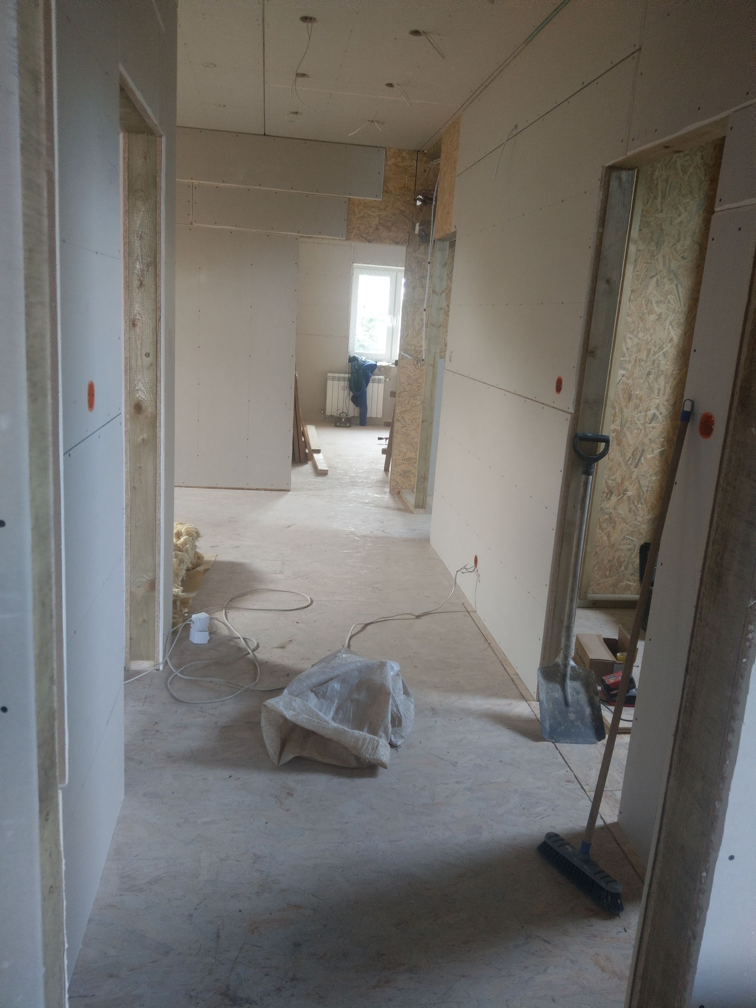 Korytarz w trakcie wykańczania, widoczne płyty gipsowo-kartonowe na ścianach, niezamaskowane łączenia, widoczne instalacje elektryczne, częściowo wykończone futryny drzwiowe, worki z materiałami...