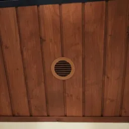 Drewniane panele ścienne w kolorze ciemnego brązu z okrągłą kratką wentylacyjną z jasnego drewna, zamontowaną centralnie.