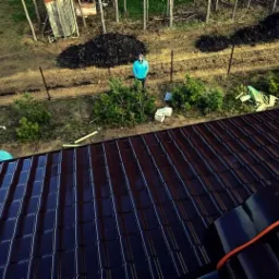 Widok z góry na nowo położony, błyszczący, brązowy dach z blachodachówki, z osobą w niebieskiej bluzie stojącą na ziemi w tle, obok ogrodzenia i sterty ziemi.