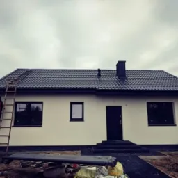 Dom jednorodzinny z ciemnym dachem z blachodachówki, widoczna drabina oparta o ścianę, pracownik w czerwonej kurtce na drabinie, pochmurne niebo w tle.