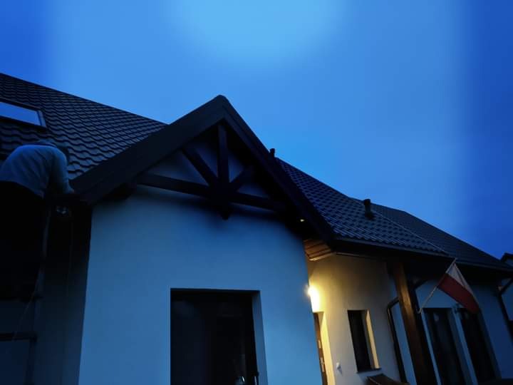 Pracownik montuje rynnę na tle ciemniejącego nieba przy nowoczesnym domu z ciemną dachówką i drewnianymi elementami wykończeniowymi.