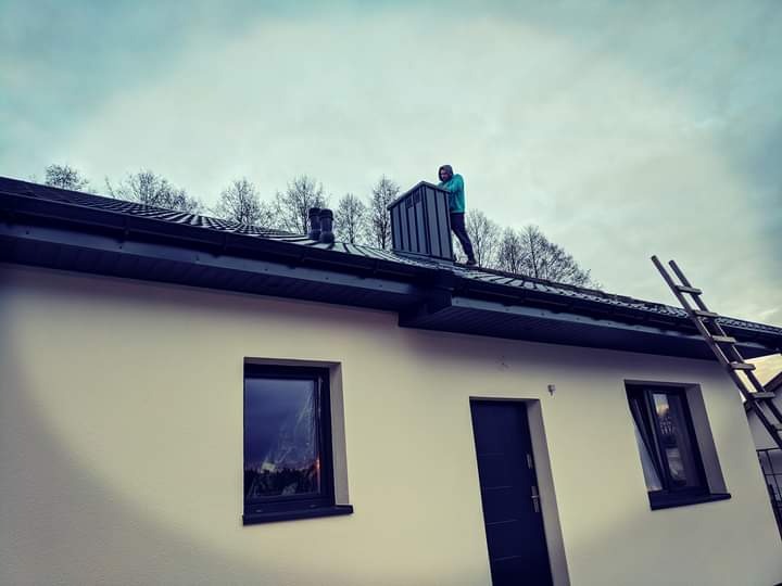 Pracownik w niebieskiej bluzie montuje metalowy komin na ciemnym dachu domu jednorodzinnego, drabina oparta o ścianę budynku.