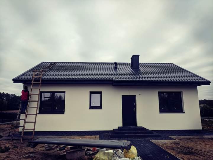 Dom jednorodzinny z ciemnym dachem z blachodachówki, widoczna drabina oparta o ścianę, pracownik w czerwonej kurtce na drabinie, pochmurne niebo w tle.