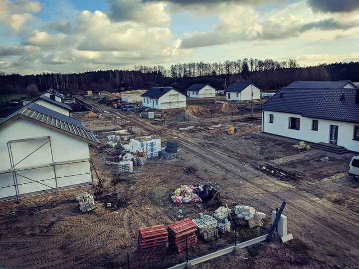 Widok z góry na plac budowy z kilkoma domami w stanie surowym zamkniętym, z ciemnymi dachami i białymi elewacjami, otoczonymi przez błotniste tereny i las w tle, pod pochmurnym niebem.