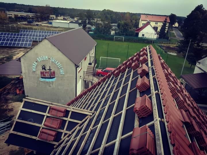 Widok z góry na konstrukcję dachu z deskami i częściowo ułożoną czerwoną dachówką, w tle budynek z napisem 'Wiara, Rozum, Świadectwo - Przystań' oraz boisko sportowe.