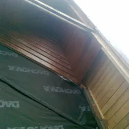 Widok z dołu na konstrukcję dachu z deskowaniem i membraną dachową z widoczną drabiną aluminiową.