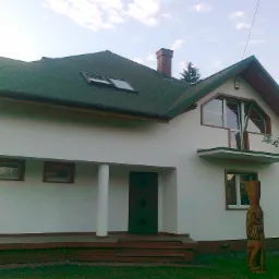 Nowo wybudowany dom jednorodzinny z zielonym dachem, brązowymi obramowaniami okien i rzeźbą drewnianą przed wejściem.