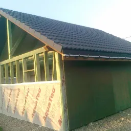 Konstrukcja szkieletowa domu w trakcie budowy, widoczne drewniane ramy, czarna dachówka i folia ochronna AquaTec na ścianie. Częściowo obita płyta OSB.