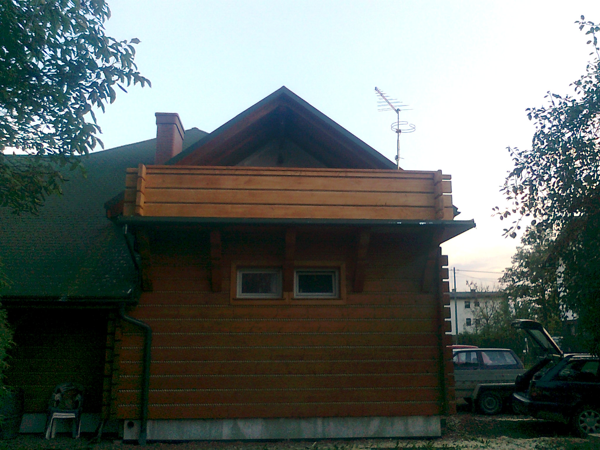 Fragment ściany domu drewnianego z balkonem, widoczne dwa okna, rynna spustowa, antena na dachu i zaparkowane samochody w tle.