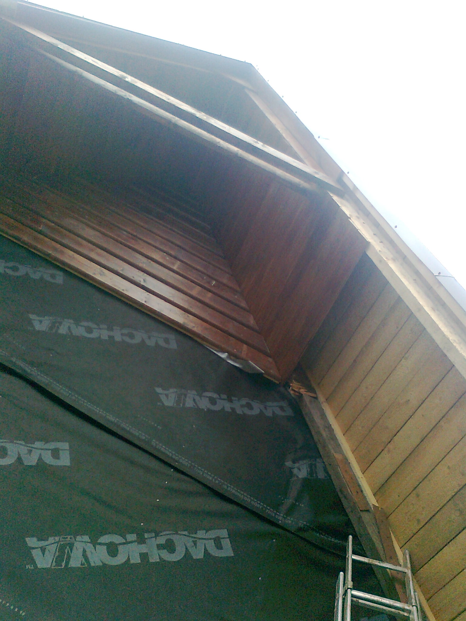 Widok z dołu na konstrukcję dachu z deskowaniem i membraną dachową z widoczną drabiną aluminiową.