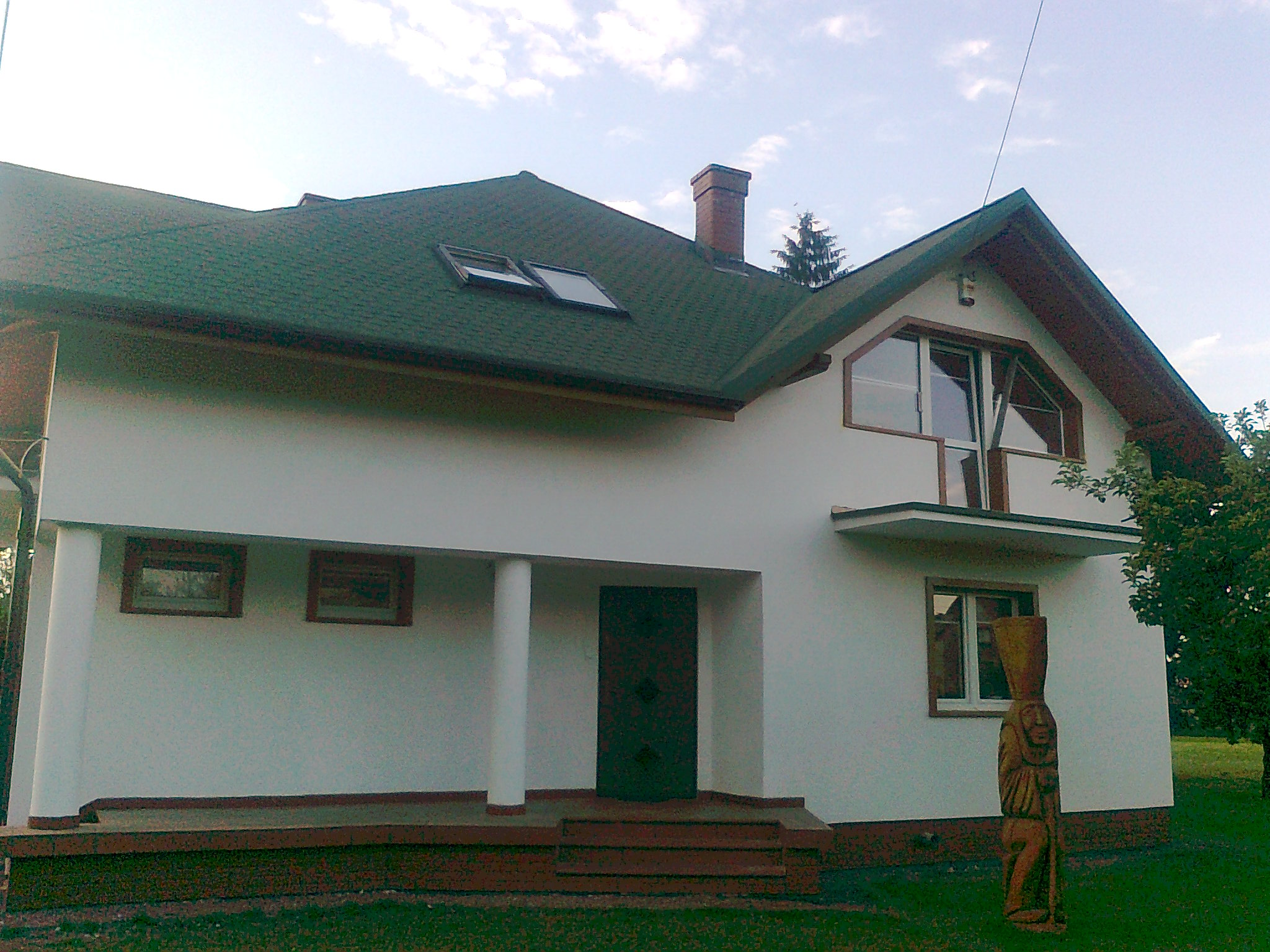 Nowo wybudowany dom jednorodzinny z zielonym dachem, brązowymi obramowaniami okien i rzeźbą drewnianą przed wejściem.