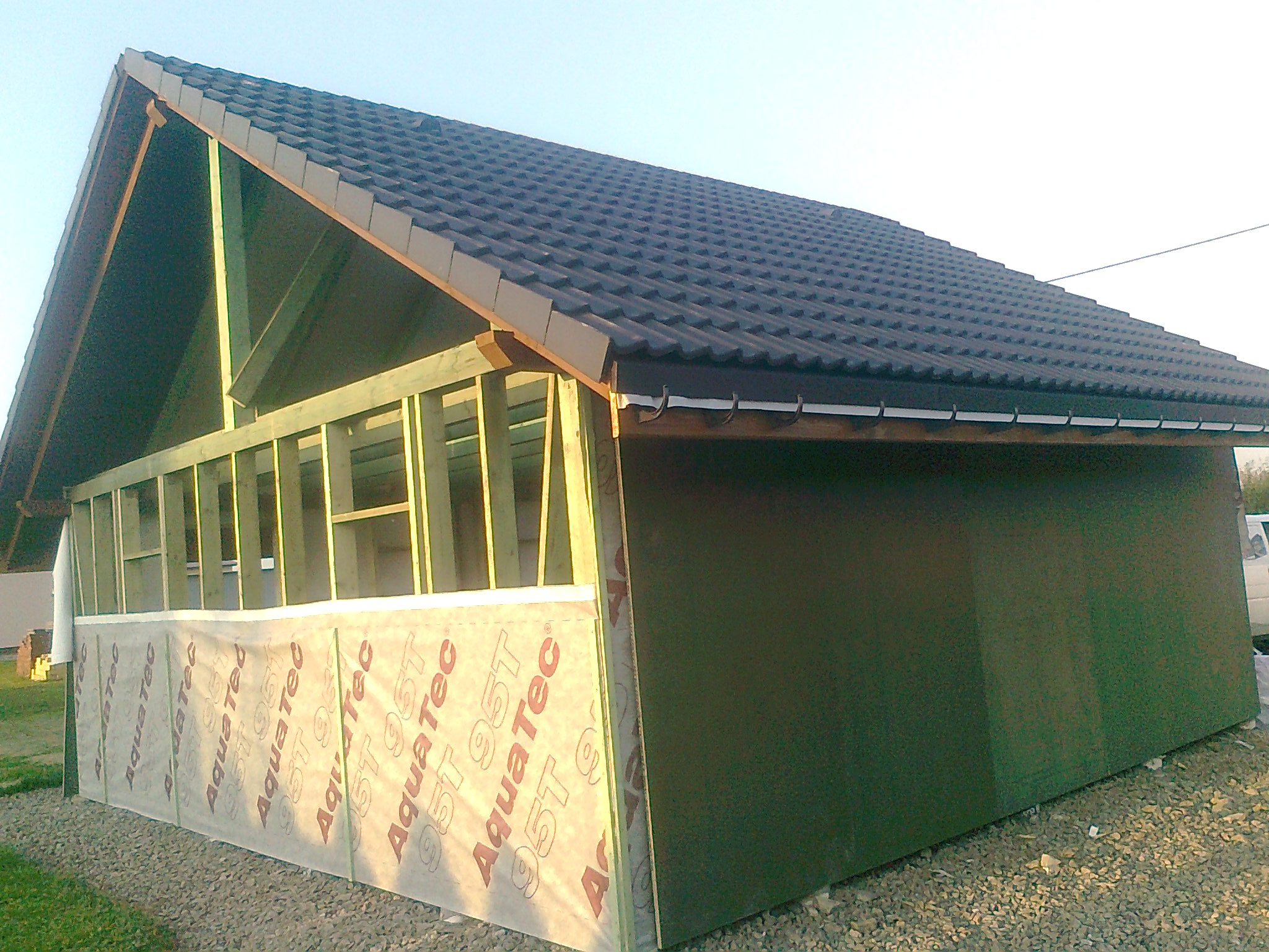 Konstrukcja szkieletowa domu w trakcie budowy, widoczne drewniane ramy, czarna dachówka i folia ochronna AquaTec na ścianie. Częściowo obita płyta OSB.