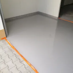 Wylewka samopoziomująca w szarym odcieniu, zabezpieczona pomarańczową taśmą, wykonana w garażu w Siedlcach, z widocznym fragmentem kostki brukowej przy wejściu.