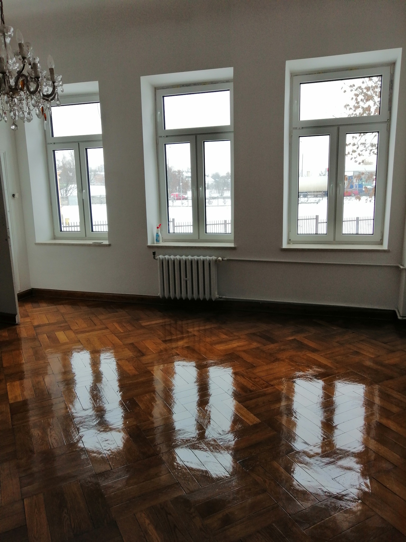Błyszcząca, odnowiona podłoga parkietowa w przestronnym pokoju z trzema oknami, przez które widać zimowy krajobraz; na parapecie butelka środka czyszczącego, a z sufitu zwisa kryształowy żyrandol.