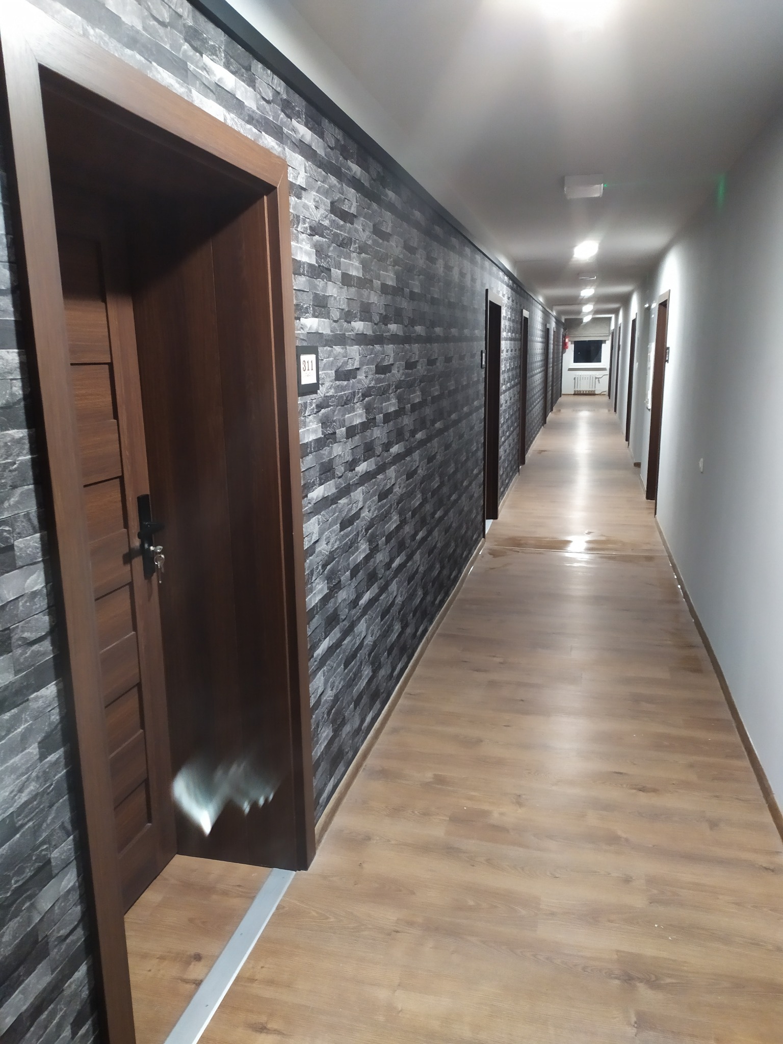 Długi korytarz hotelowy z panelami podłogowymi, ściana wykończona imitacją kamienia, widoczne drzwi do pokoi z numerami.