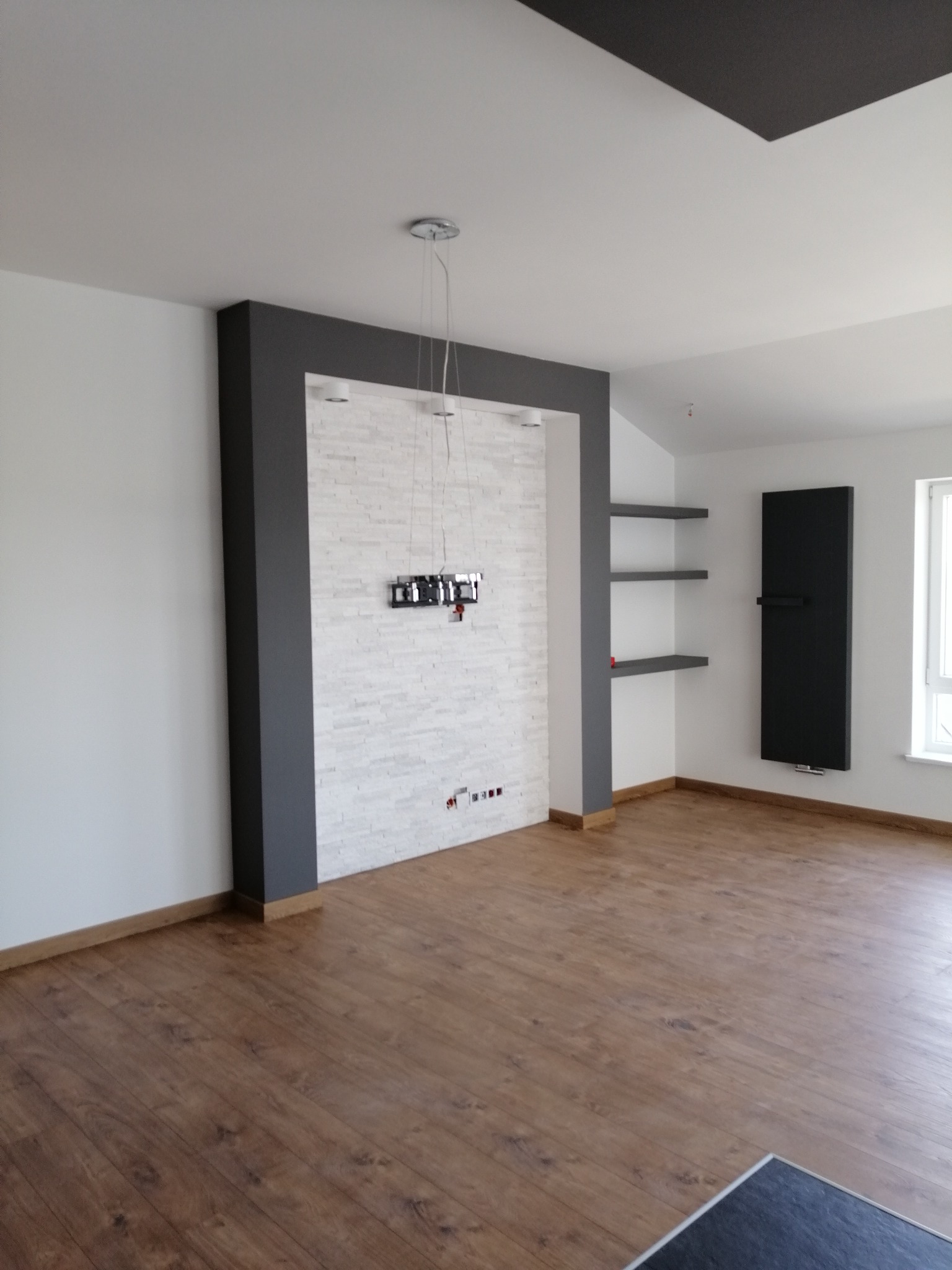 Nowoczesne wnętrze z drewnianą podłogą, ścianą pokrytą białymi płytkami imitującymi cegłę, obudowaną szarą ramą, wiszącym uchwytem na telewizor i minimalistycznymi półkami.