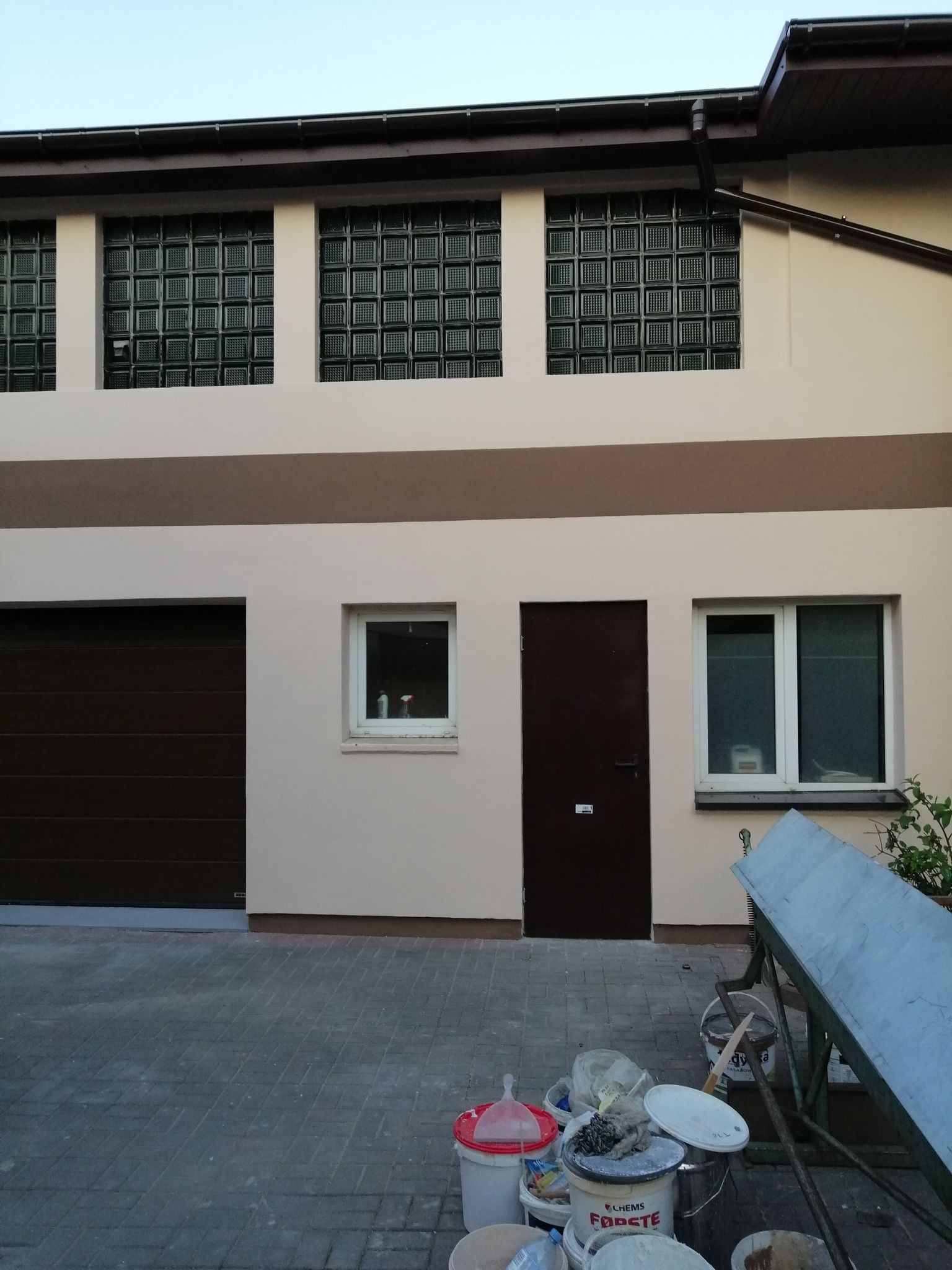 Dwukondygnacyjny budynek z oknami z luksferów, brązową bramą garażową i drzwiami, po remoncie elewacji, z widocznymi wiadrami i narzędziami malarskimi na kostce brukowej.