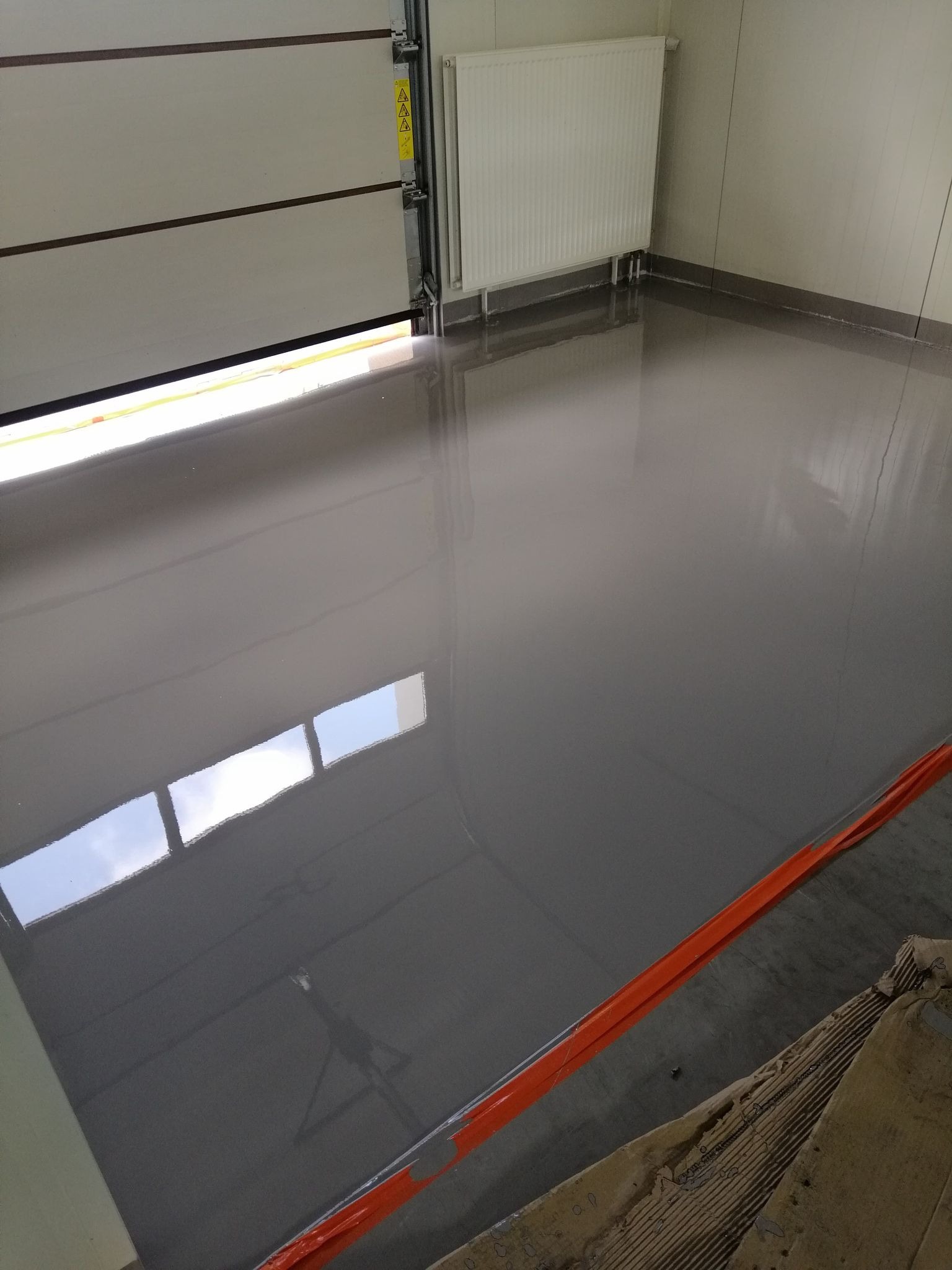 Świeżo wylana, szara posadzka przemysłowa z żywicy epoksydowej w hali, z widocznym odbiciem okien w lśniącej powierzchni, zabezpieczona pomarańczową taśmą na krawędziach i z częściowo otwartą bramą...
