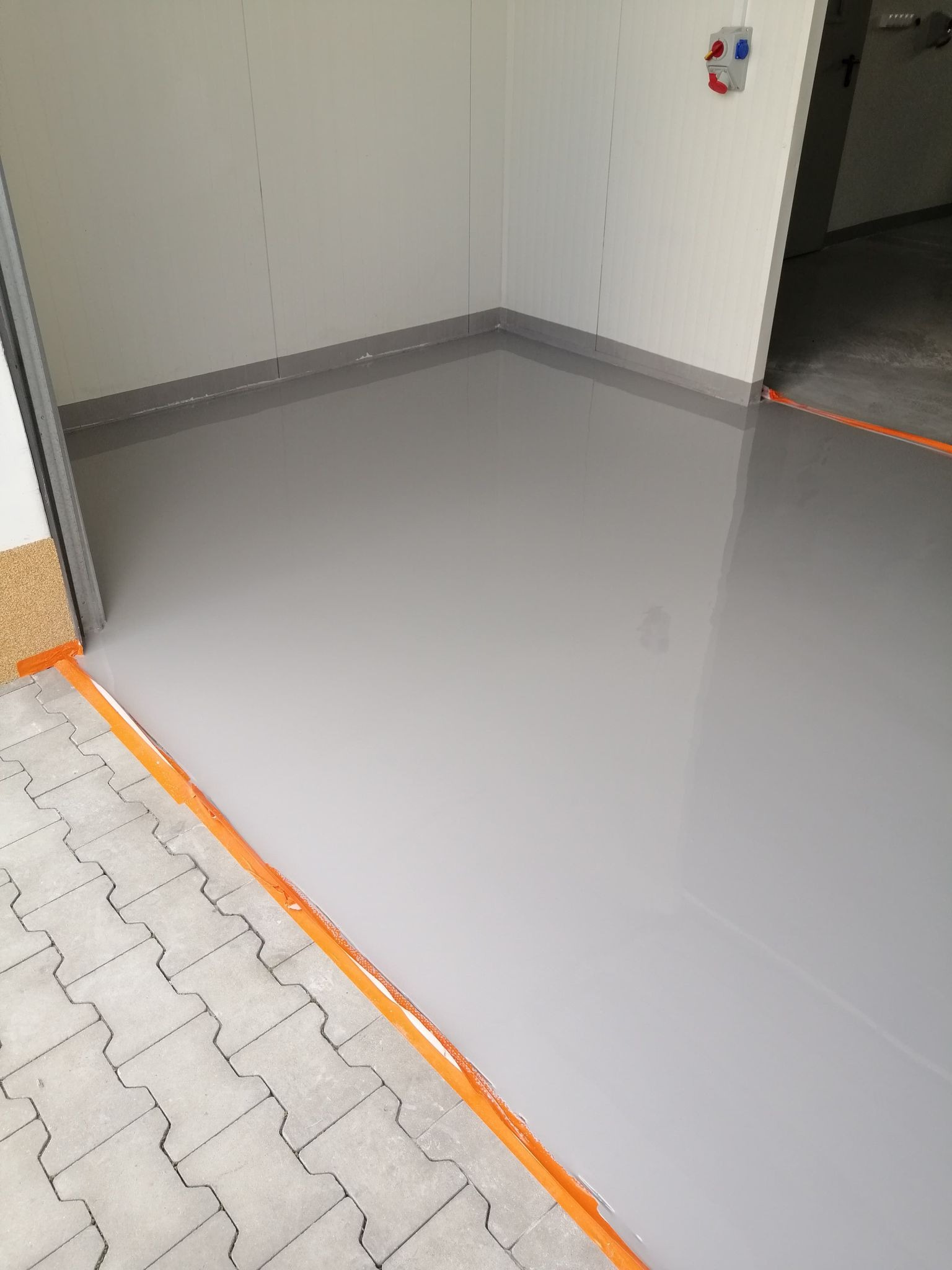 Wylewka samopoziomująca w szarym odcieniu, zabezpieczona pomarańczową taśmą, wykonana w garażu w Siedlcach, z widocznym fragmentem kostki brukowej przy wejściu.