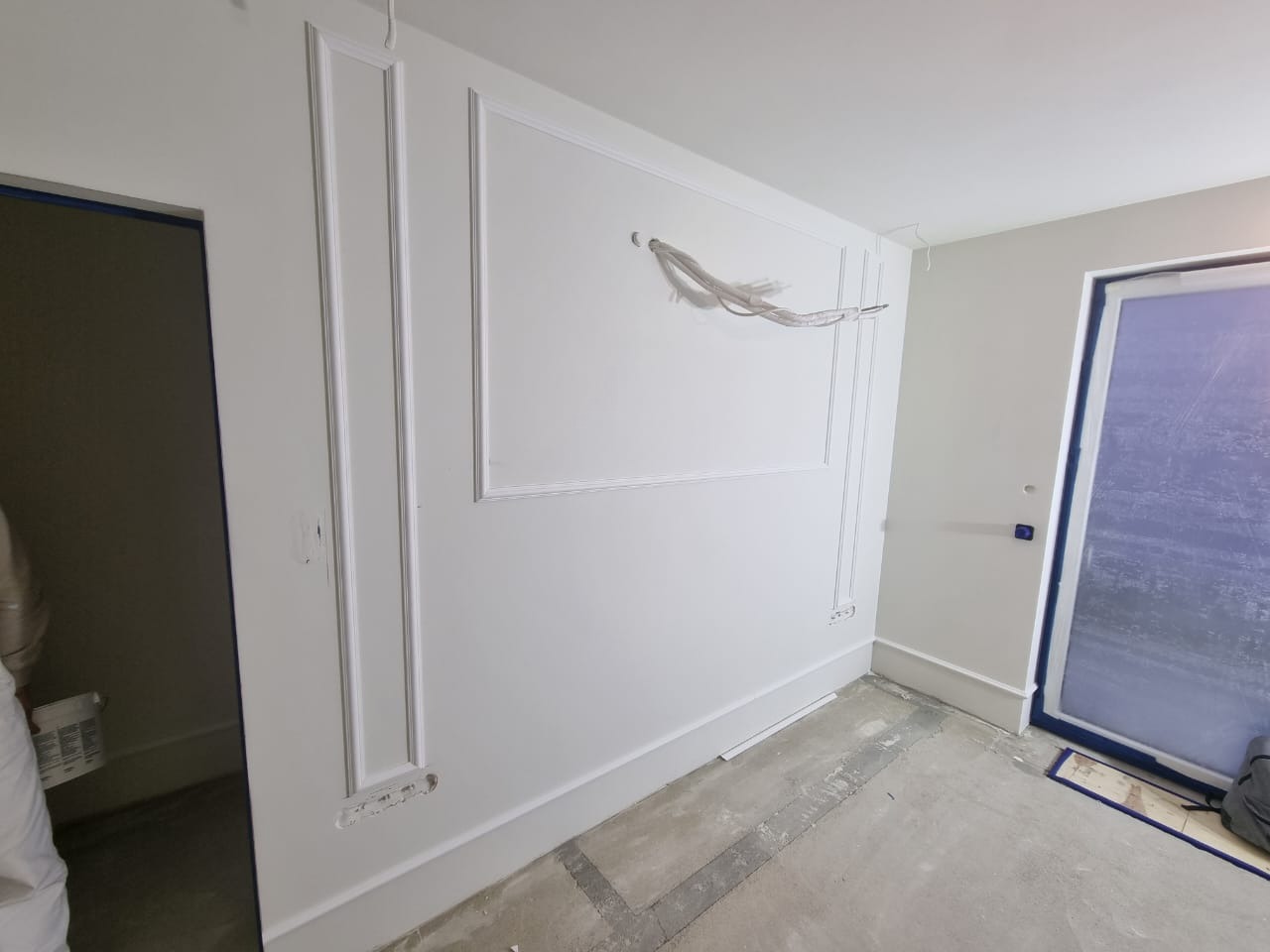 Białe ściany z dekoracyjnymi listwami i ramkami w trakcie montażu, widoczne wystające przewody elektryczne, surowa podłoga przed wykończeniem, zabezpieczone okno.