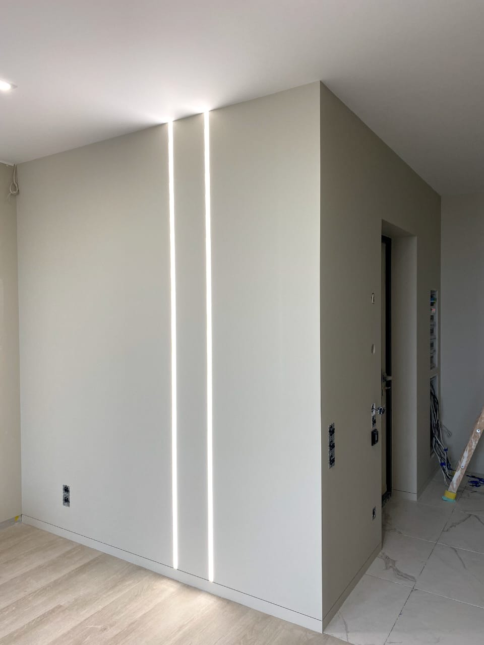 Minimalistyczne wnętrze z dwiema pionowymi liniami światła LED w ścianie, szara kolorystyka, parkiet i płytki podłogowe, widoczne puszki elektryczne i fragment drabiny.