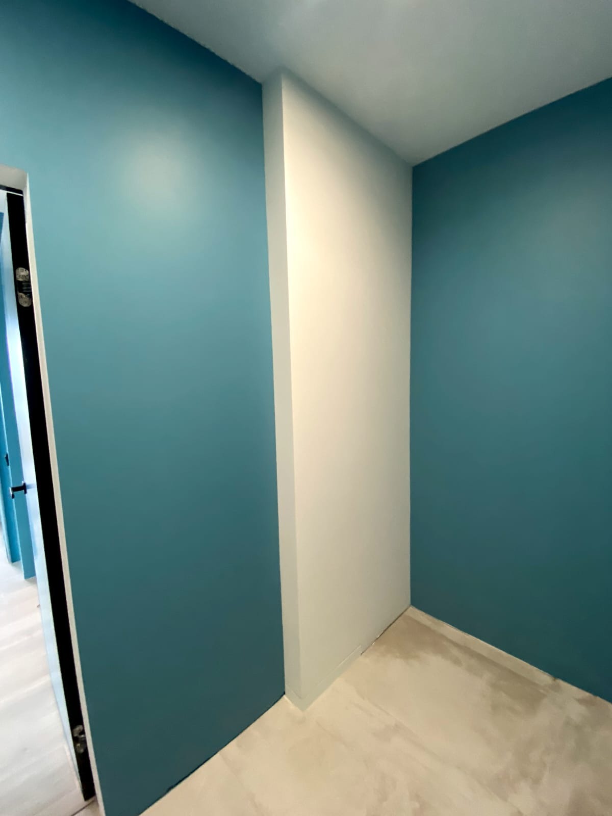 Świeżo pomalowane na turkusowo ściany w rogu pokoju, z białą kolumną i podłogą w jasnym odcieniu beżu, widok na korytarz z otwartymi drzwiami.